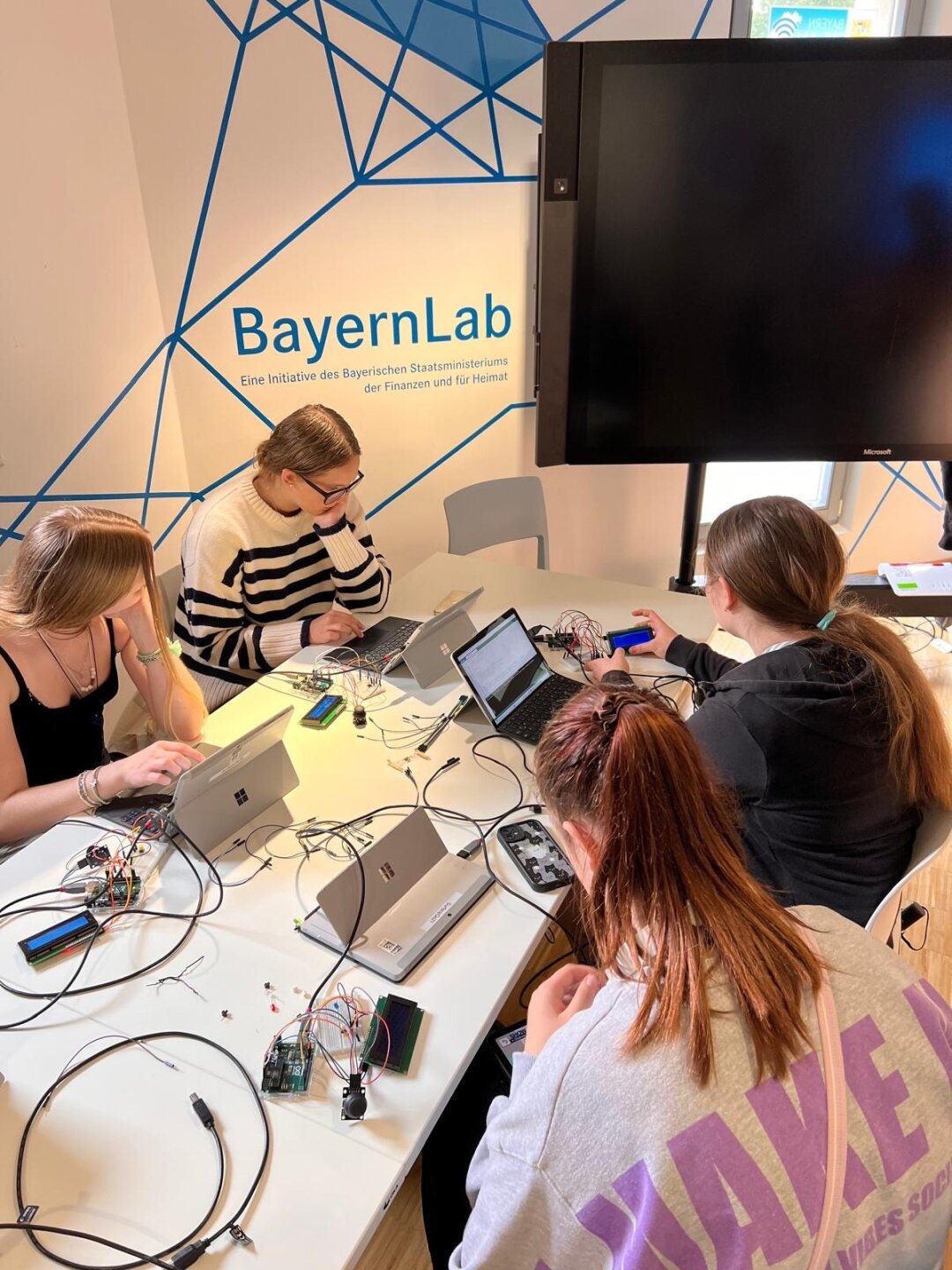 4 junge Frauen sitzen an Laptops an einem Tisch mit vielen Kabeln. Im Hintergrund der Schriftzug 